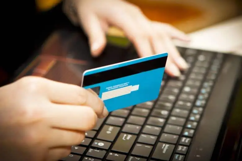 Mobile Shopping Safety For The Holiday Season 3 hands holding a blue card above a laptop, showcasing online shopping convenience this holiday season.