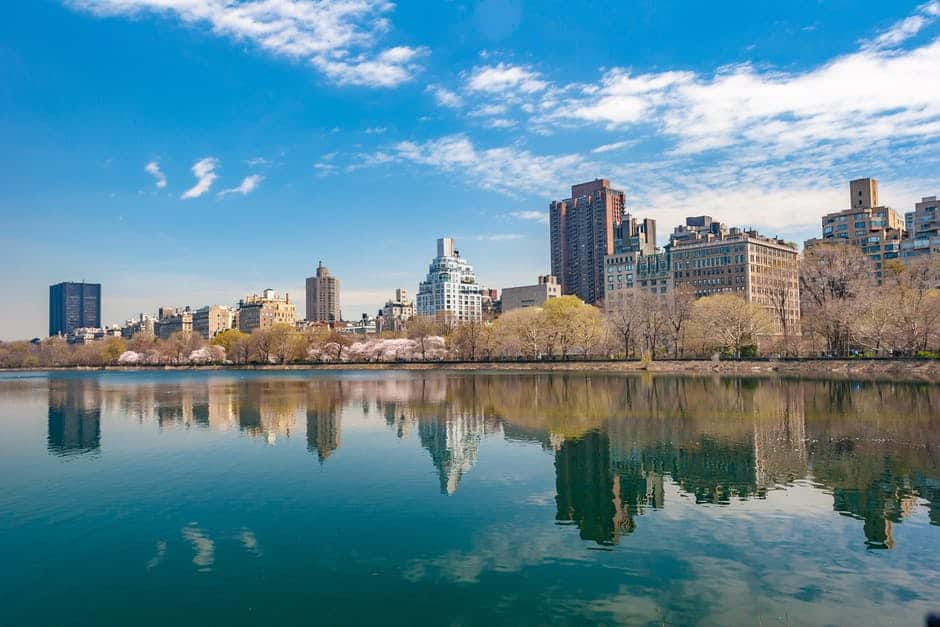 city skyline reflected in calm water under a clear blue sky and fluffy clouds.