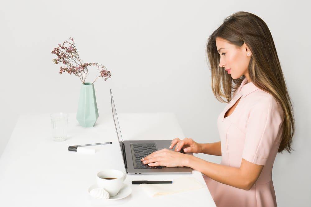 young woman focused on laptop at minimalistic white desk with coffee and notepad.