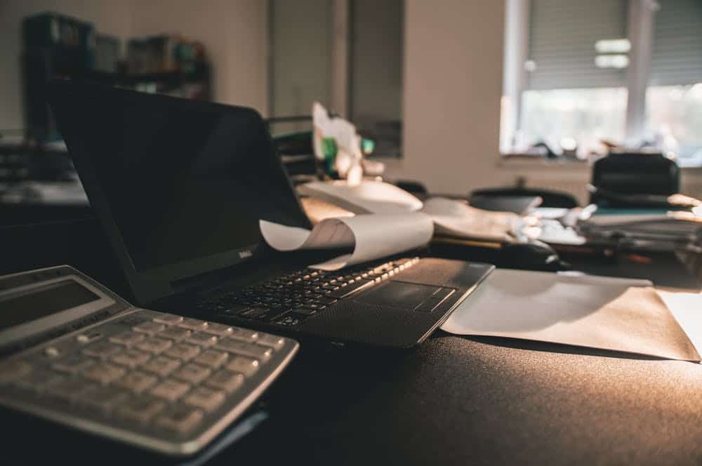 a busy office workspace with a laptop, calculator, and scattered papers under warm natural light.