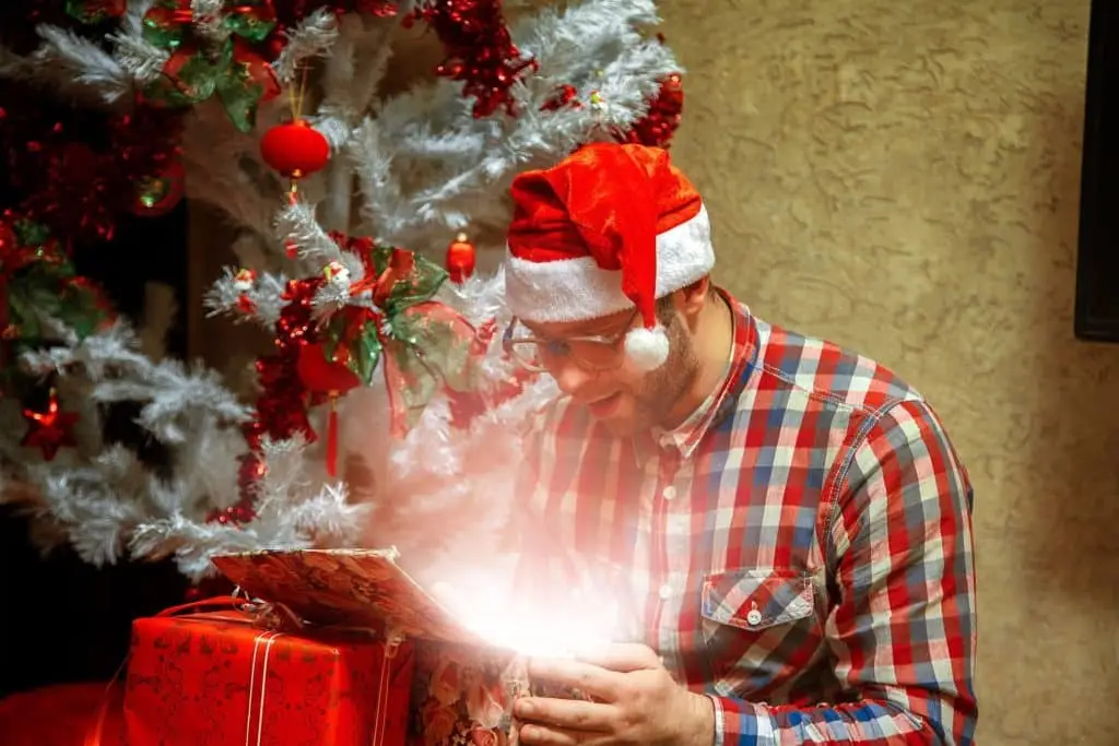 man in a santa hat excitedly opens a glowing gift under a festive christmas tree.