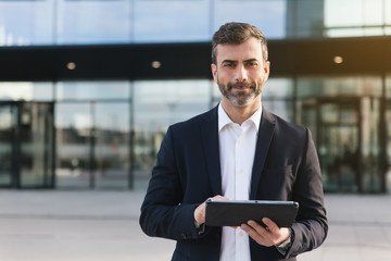 confident technology director in tailored suit holding tablet outside modern glass building.