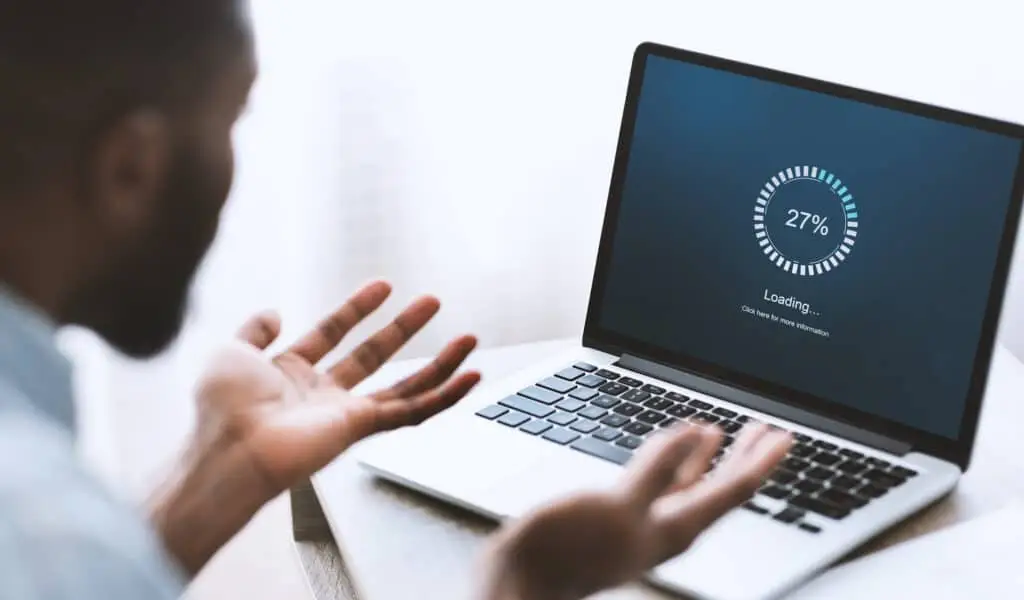 3 Simple Problems That Cause A Slow Computer 2 frustrated man staring at laptop loading screen in professional workspace, hands raised in disbelief.