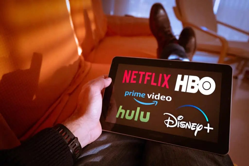 Streaming Technology And How It Works 5 man relaxing with tablet showing logos of streaming services netflix, hulu, hbo, amazon, and disney+.