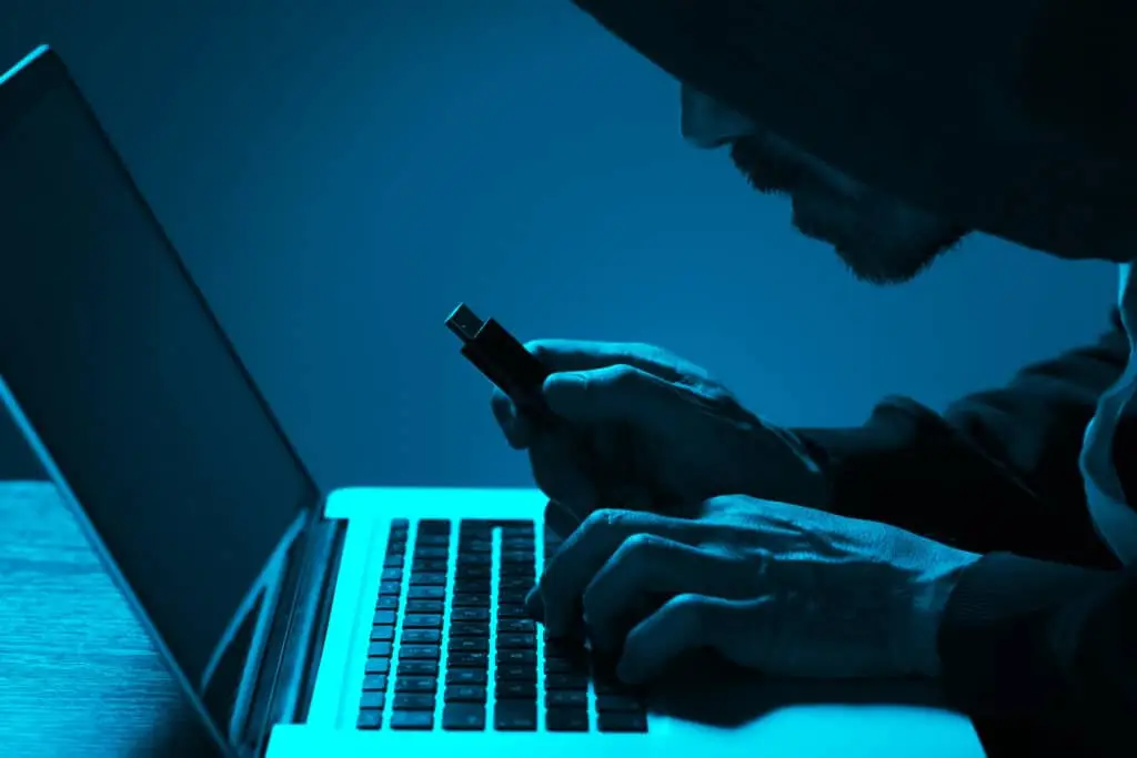 The Raspberry Robin Worm 5 hooded hacker stealing data from a laptop in a dimly lit room with usb drive.